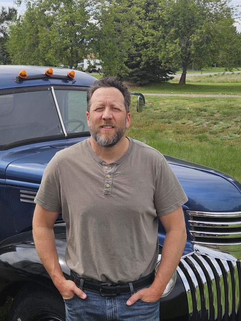 Brian Heck standing beside a vintage blue pickup truck in a rural Montana setting